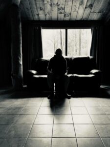 Photo of Dave sitting in a dark room. Light is coming in from the rear window through which you can see forest. The roof is made of wood and the floor of tiles.