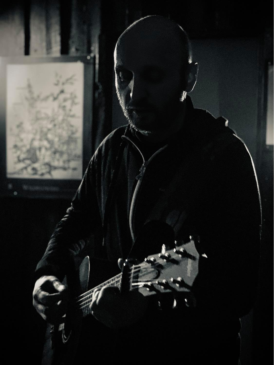 Photo of Dave standing in dark room and strumming a guitar. The guitar is catching the light as well as the stubble on one side of Dave's face.